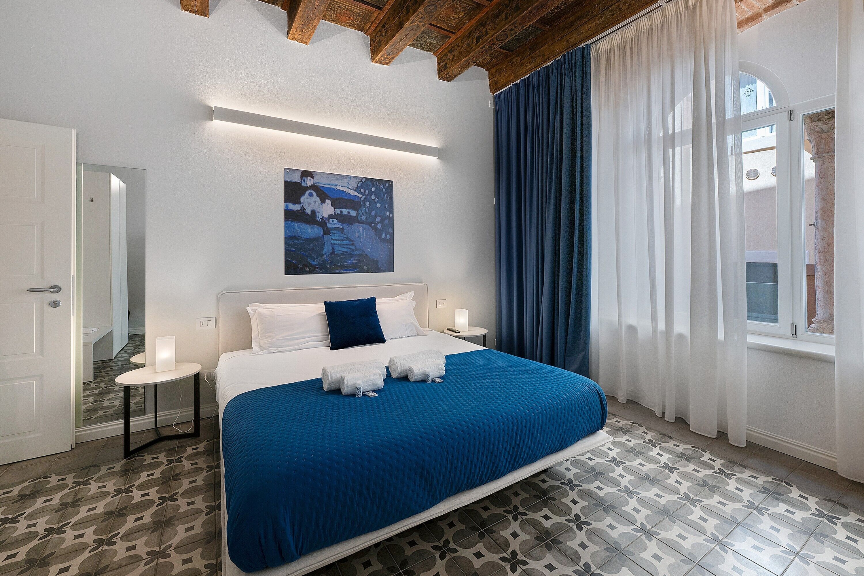 A bedroom with a large bed, a bedside table with a lamp, a painting on the wall, and a window with curtains.
