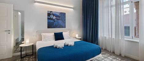 A bedroom with a large bed, a bedside table with a lamp, a painting on the wall, and a window with curtains.