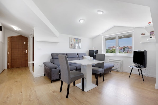 A modern living room with a grey sofa, a white dining table, and a TV on a stand.