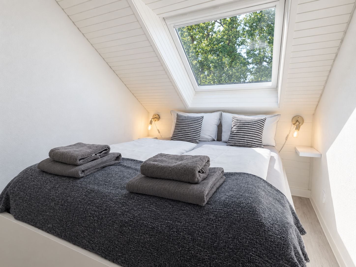 Un dormitorio con tragaluz, una cama con colcha gris texturizada, dos lámparas de noche y una mesita de noche.