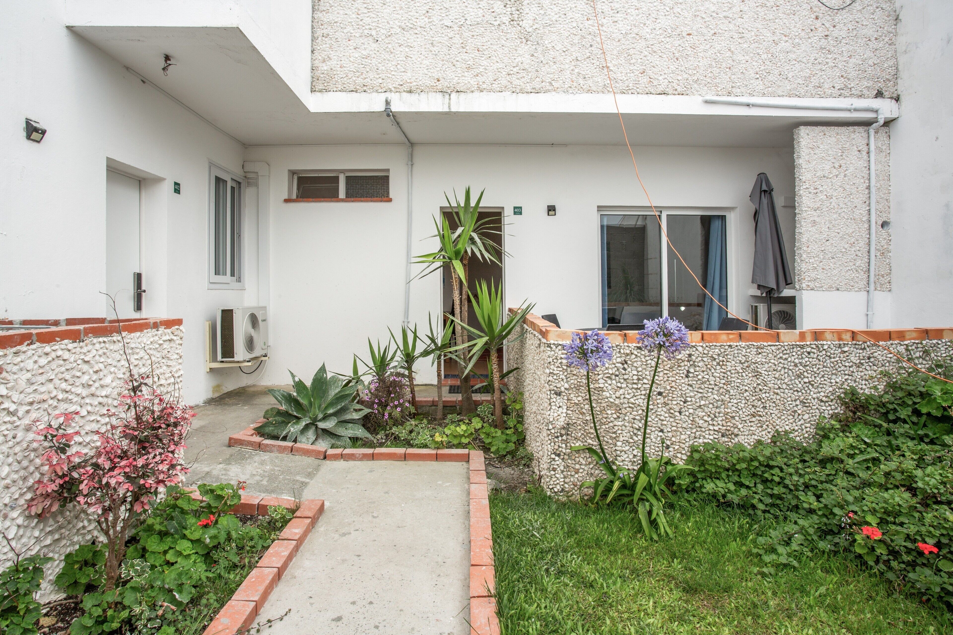 Ein Innenhof mit Betonweg, einem kleinen Garten mit verschiedenen Pflanzen, einer Tür und einem Fenster.