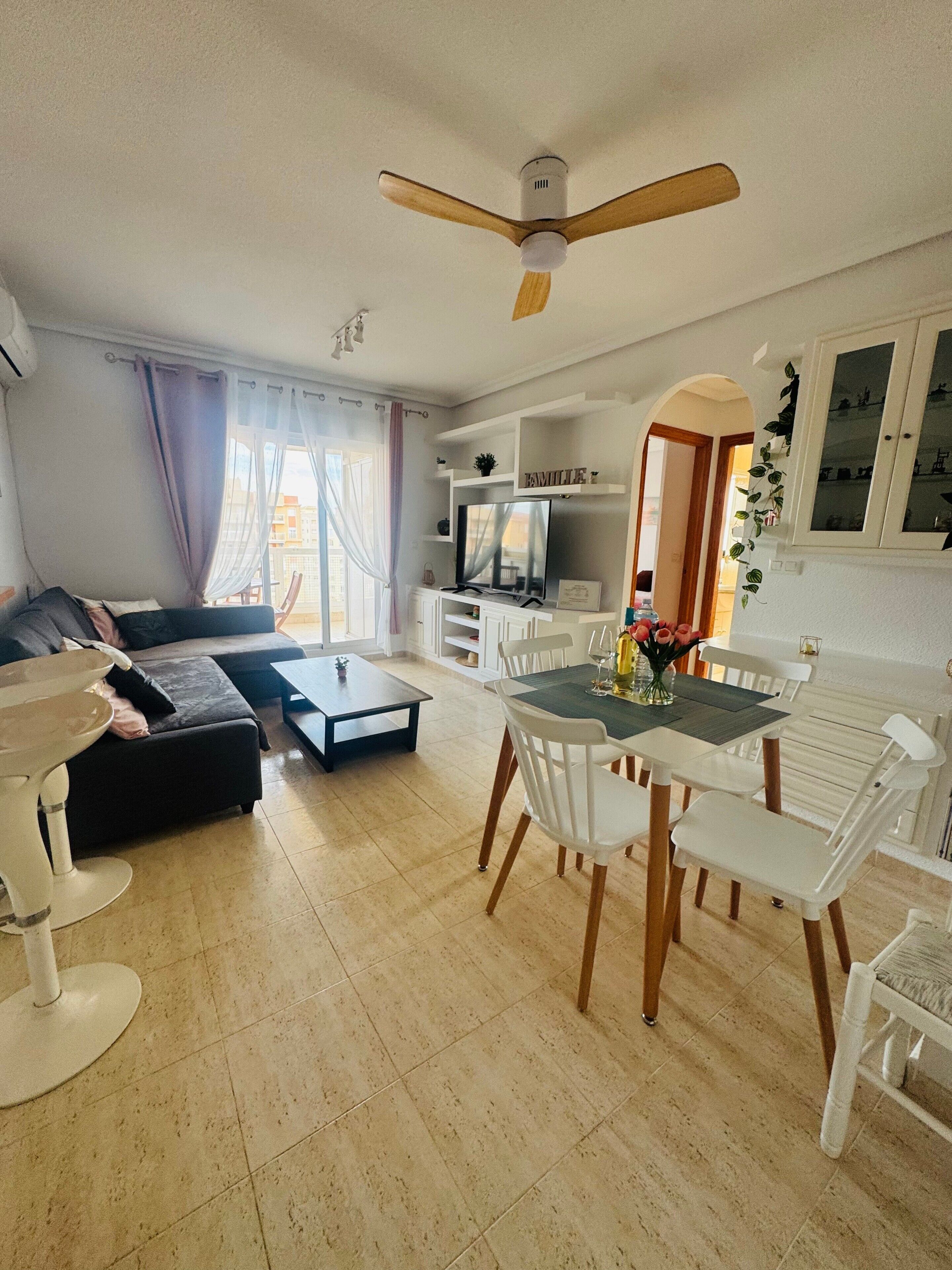 Una sala de estar con un sofá, una mesa de comedor con sillas, un ventilador de techo y un televisor sobre un mueble.