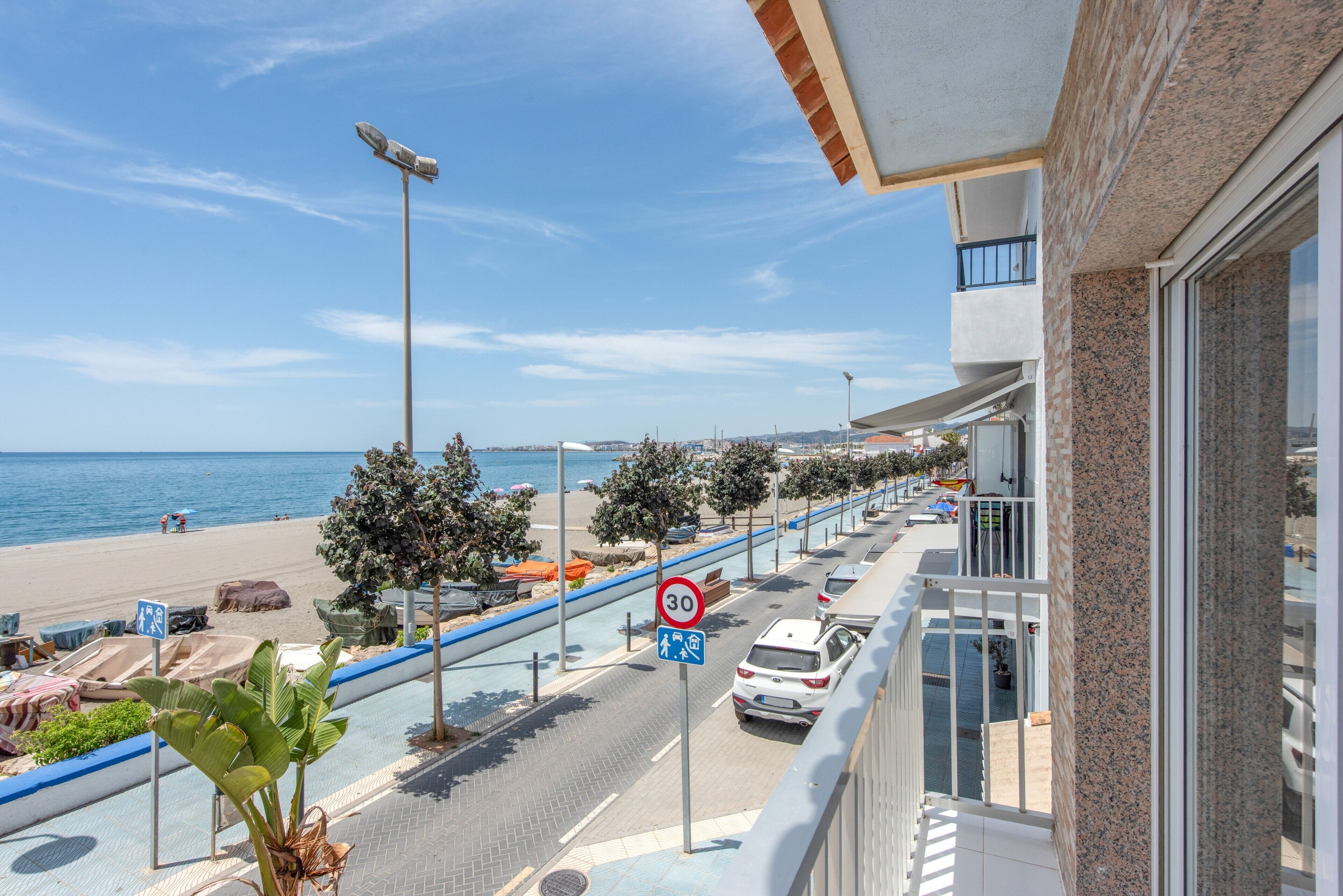 Un balcón con vista a una playa, una calle con coches estacionados y un sendero para peatones.