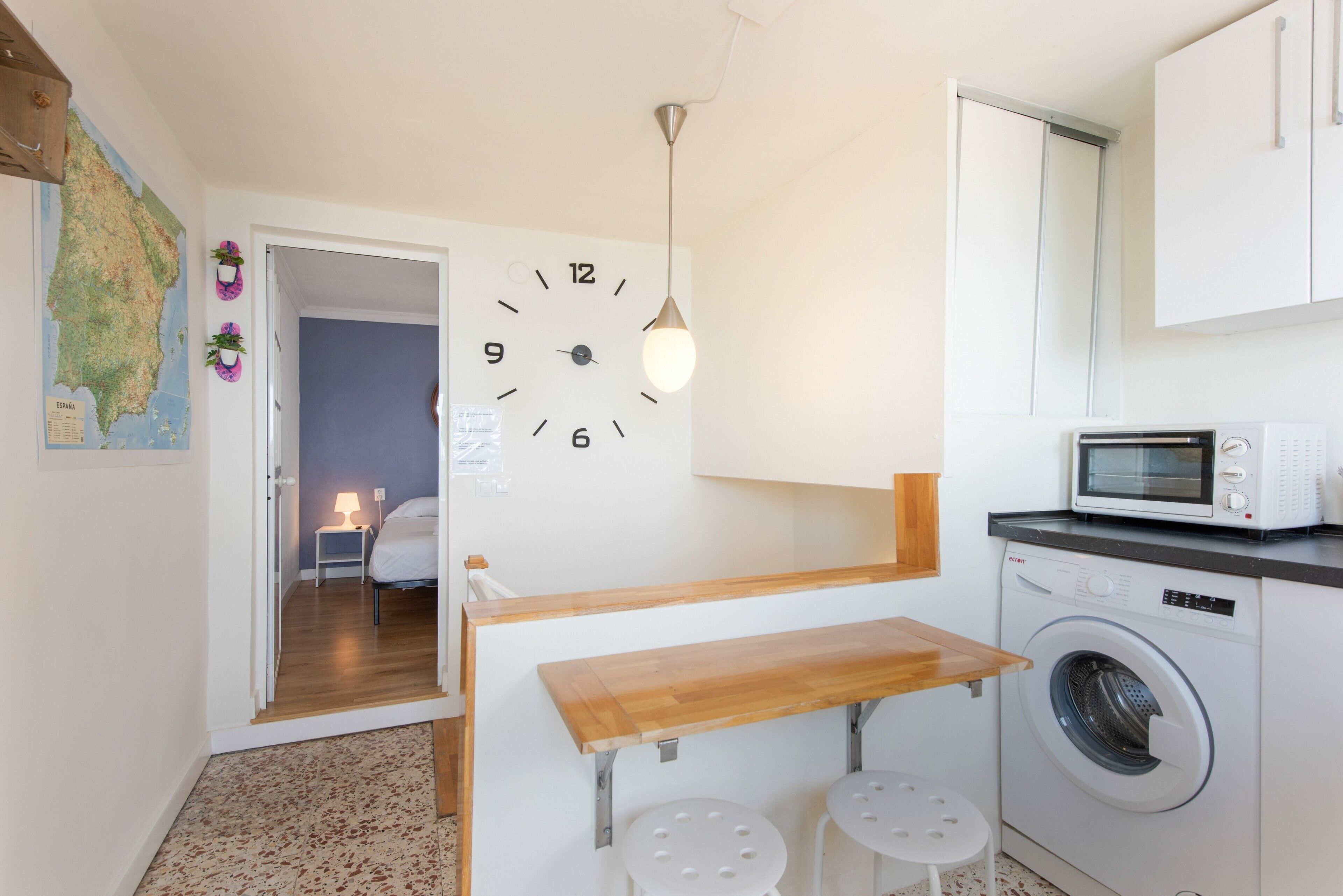 Una cocina compacta con lavadora, microondas, una mesa de madera y un reloj de pared.