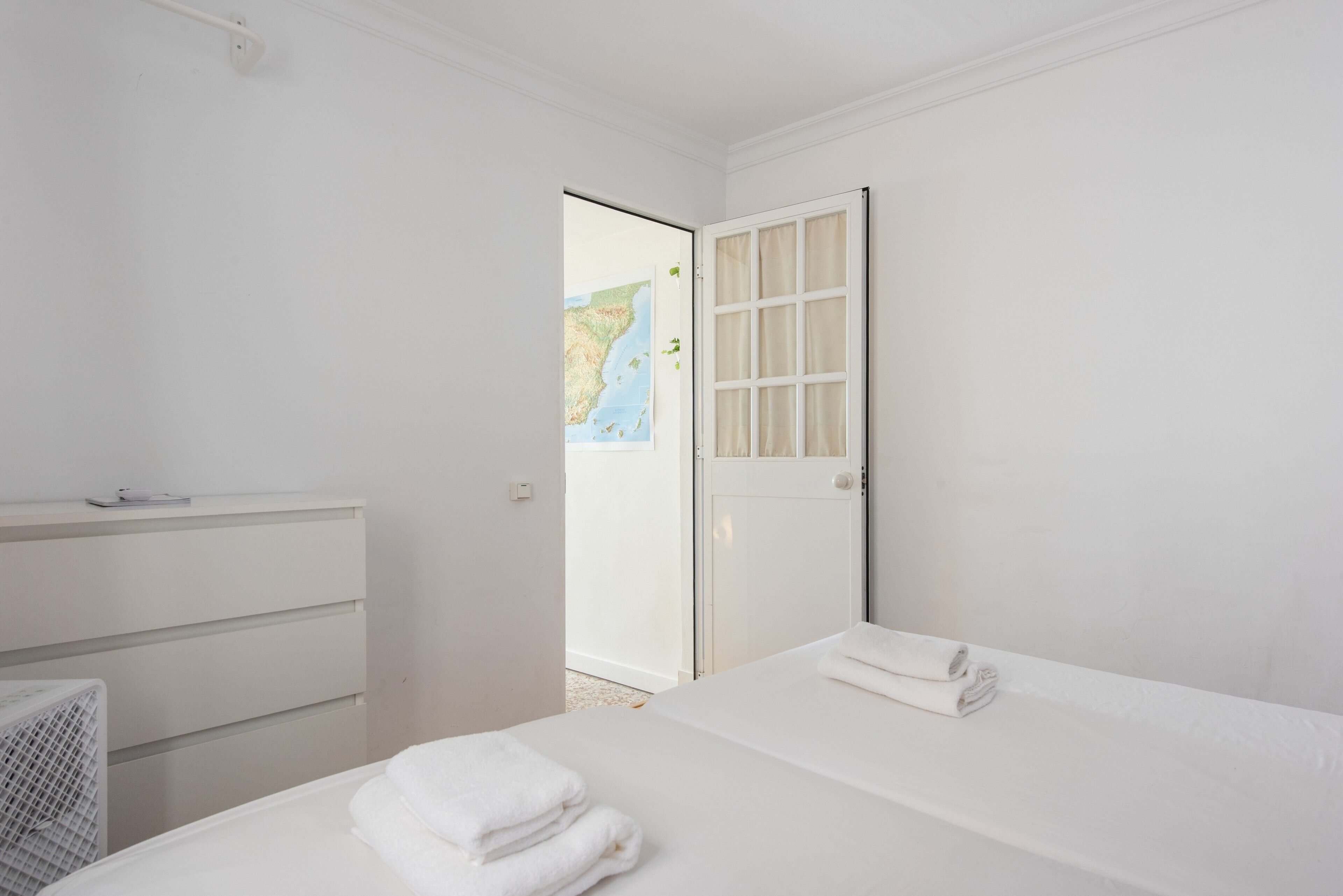 A minimalist hotel room with a bed, a small bedside table, a white dresser, and a door leading to a balcony with a map.
