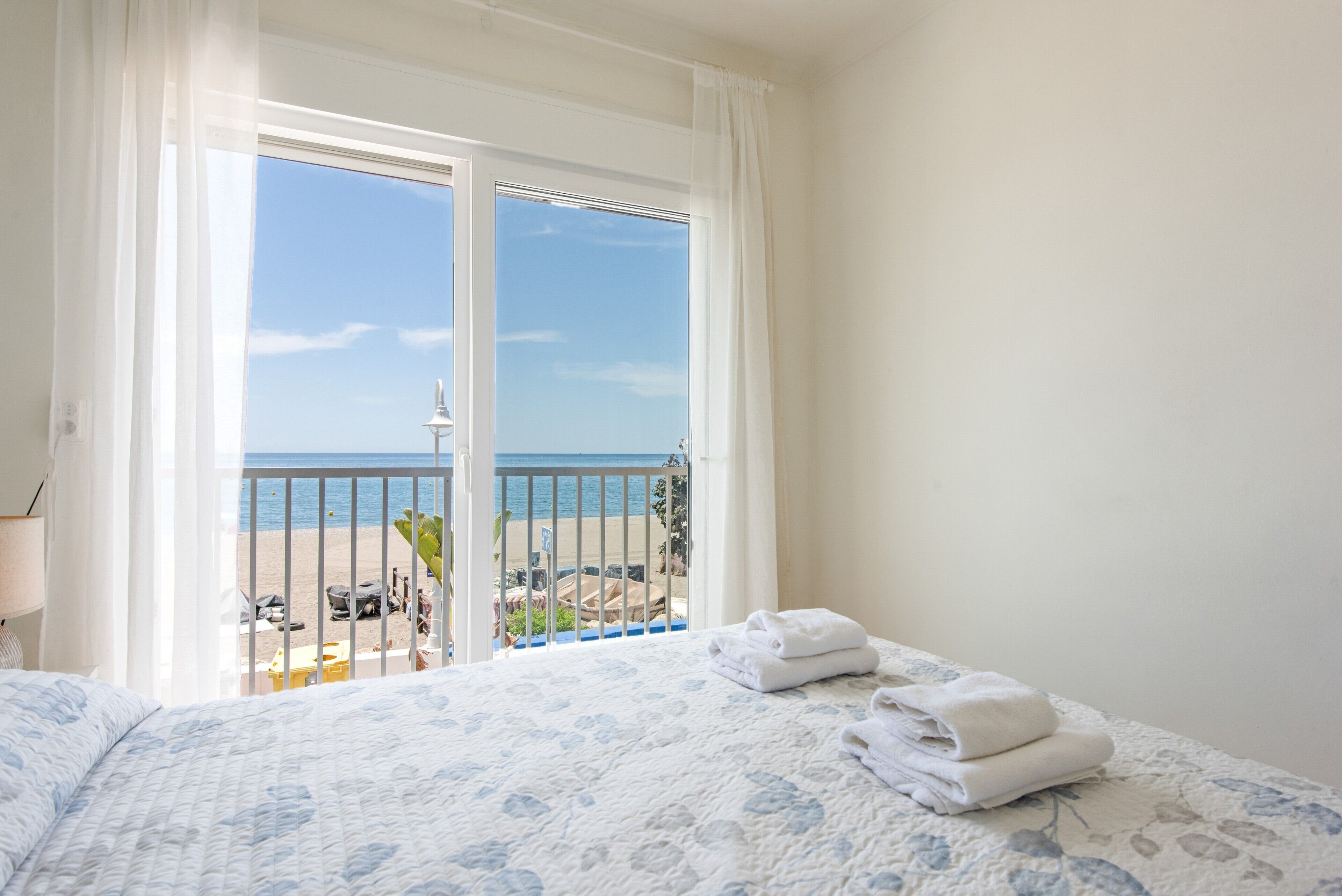 A hotel room with a bed, a balcony with a view of the beach and sea, and a window with white curtains.
