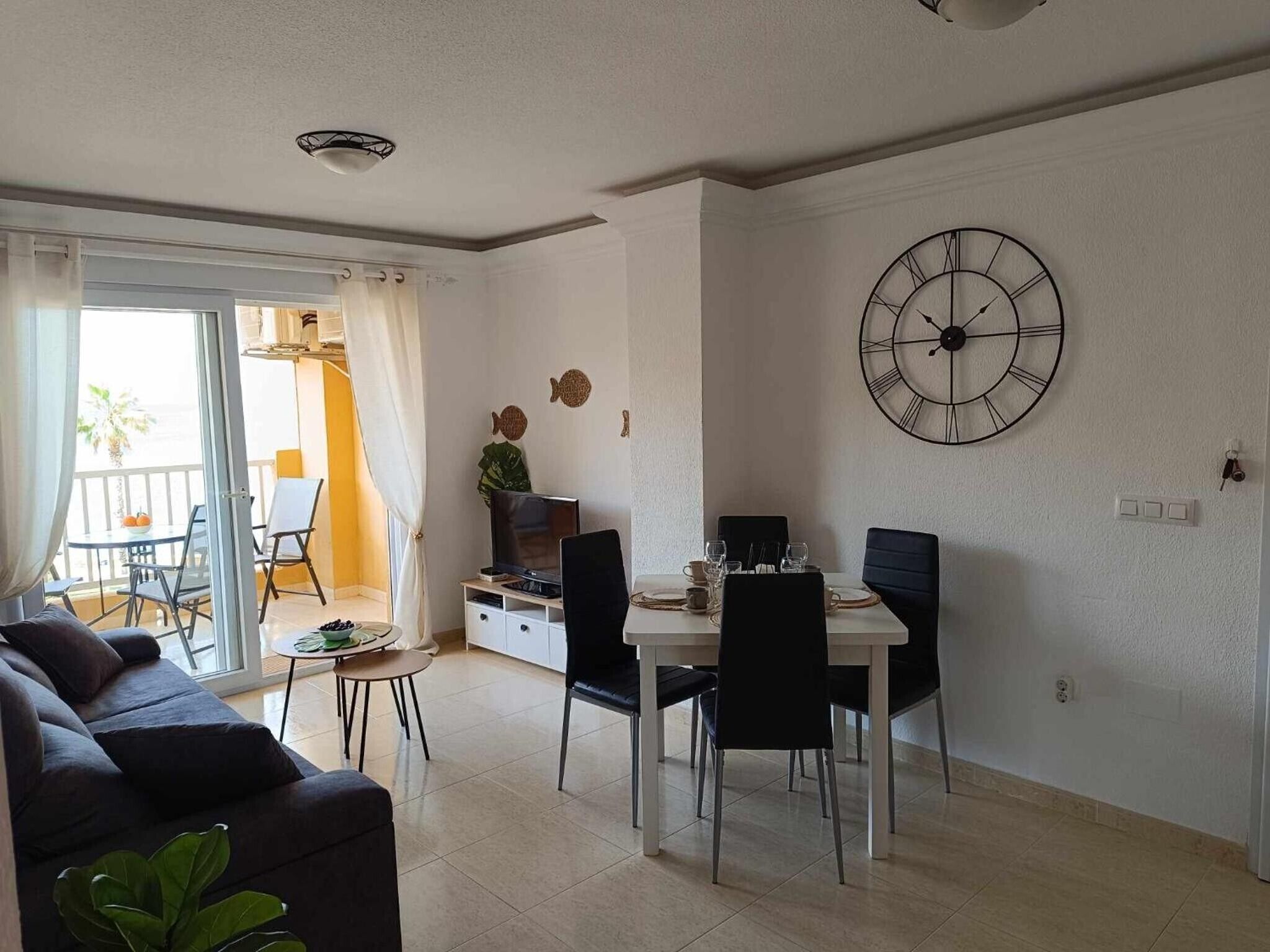 Ein modernes Wohnzimmer mit einer Couch, einem Esstisch und Stühlen. Ein Fernseher steht auf einem Schrank, eine Uhr hängt an der Wand und ein Fenster mit Vorhängen ist zu sehen.