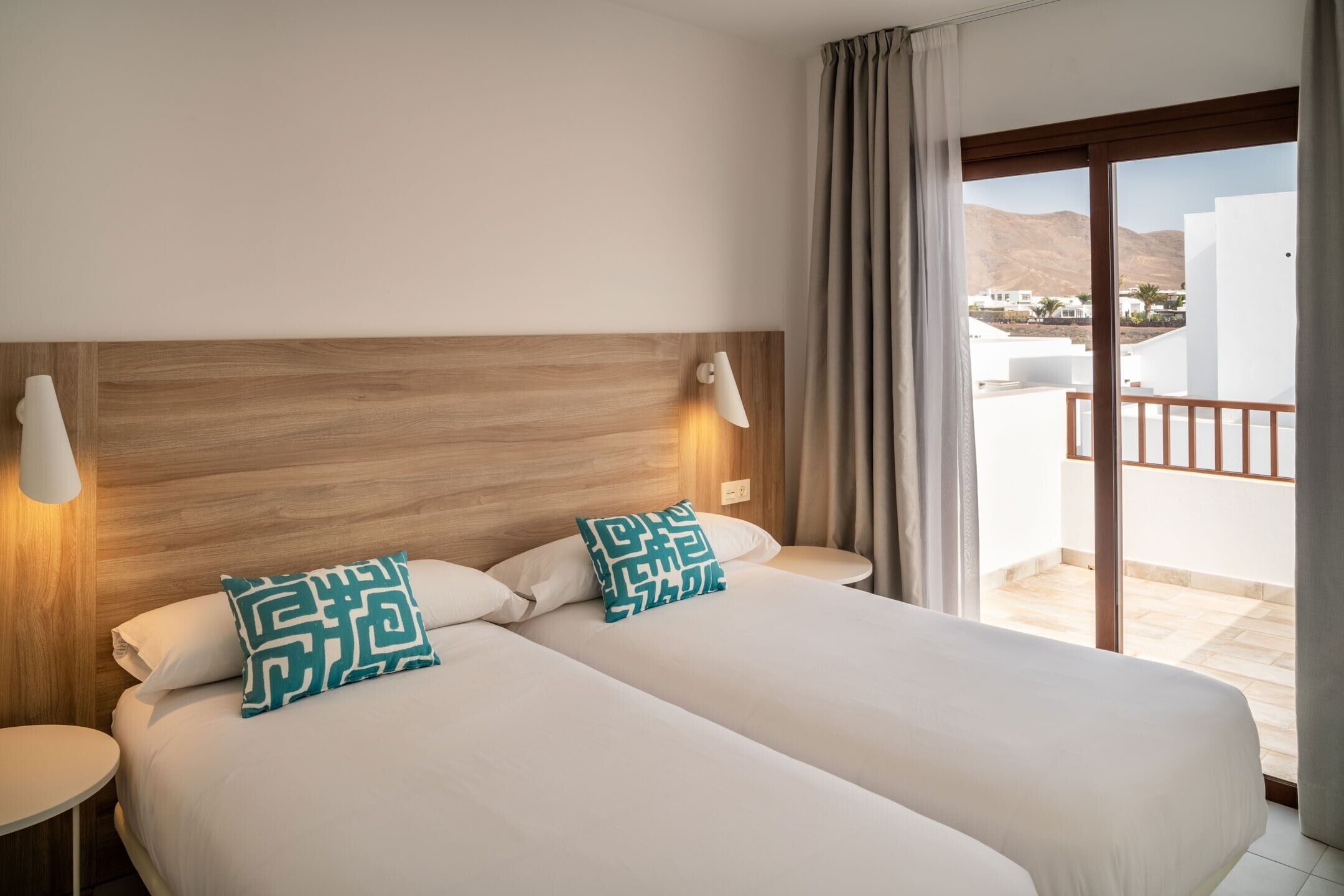 A hotel room with two beds, a wooden headboard, a balcony with a view of buildings and mountains, and a round table.