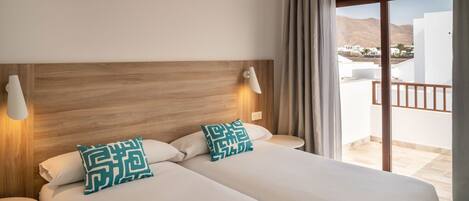 A hotel room with two beds, a wooden headboard, a balcony with a view of buildings and mountains, and a round table.