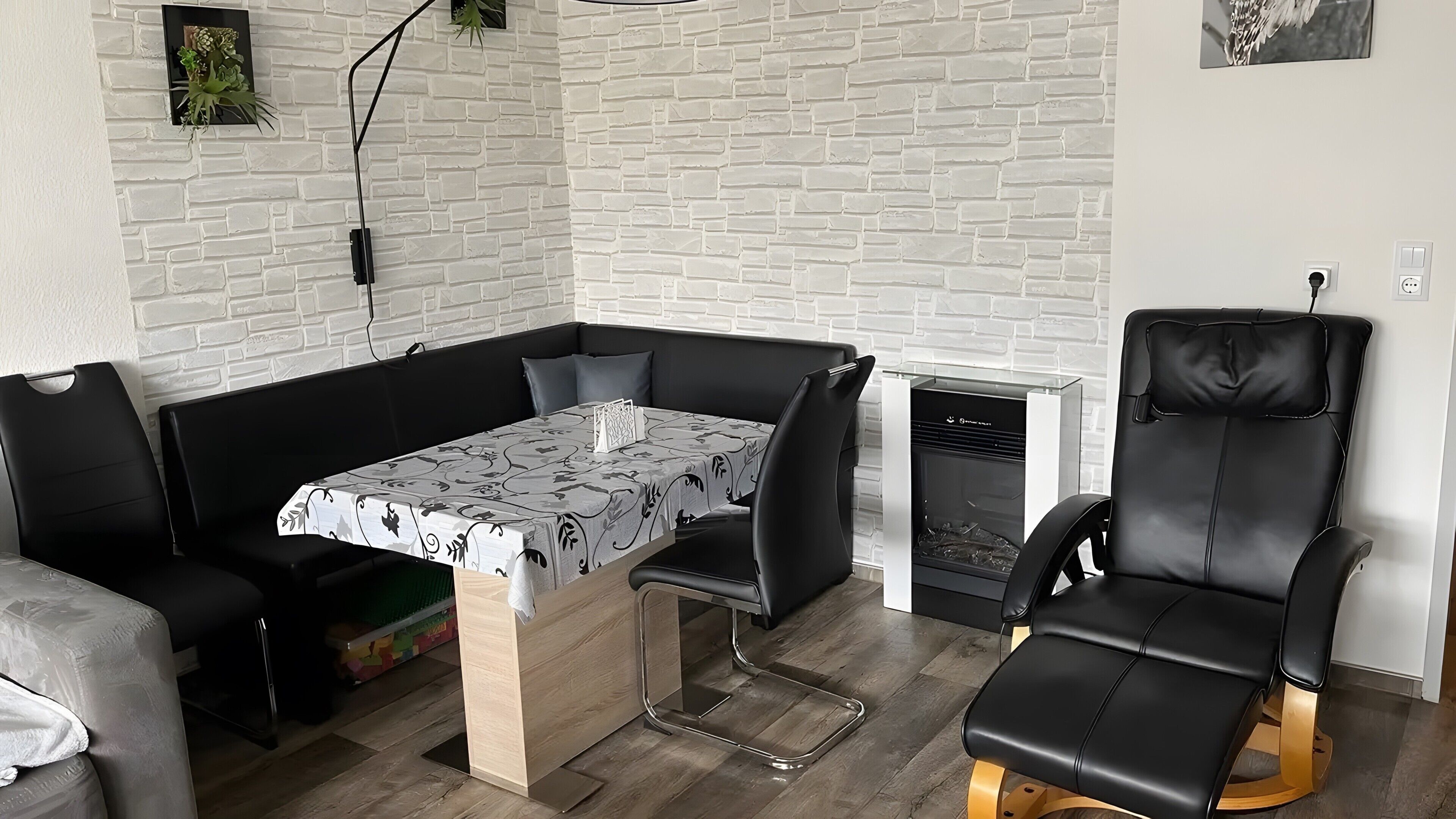 A modern living room with a white brick wall, a black leather sofa, a dining table with a patterned tablecloth, and a black leather chair.