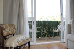 A room with a wooden chair, a sofa, and a table with chairs. There is a sliding glass door leading to an outdoor area with a hedge and a view of buildings. - Apartment 'Palma's Place' with Mountain View, Shared Garden and Wi-Fi (Caniço)