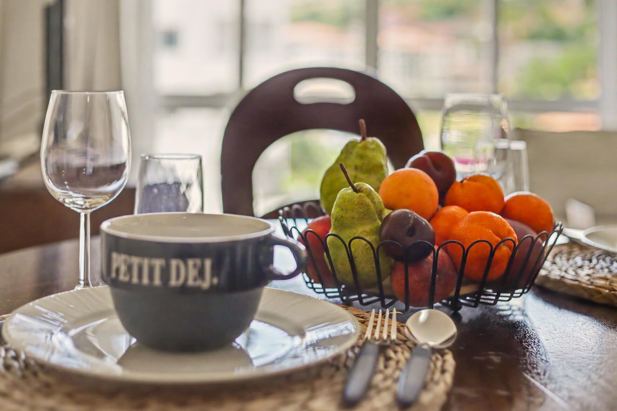 A dining table with a bowl labeled 'PETIT DEJ,' a glass of water, a fruit basket, and cutlery.