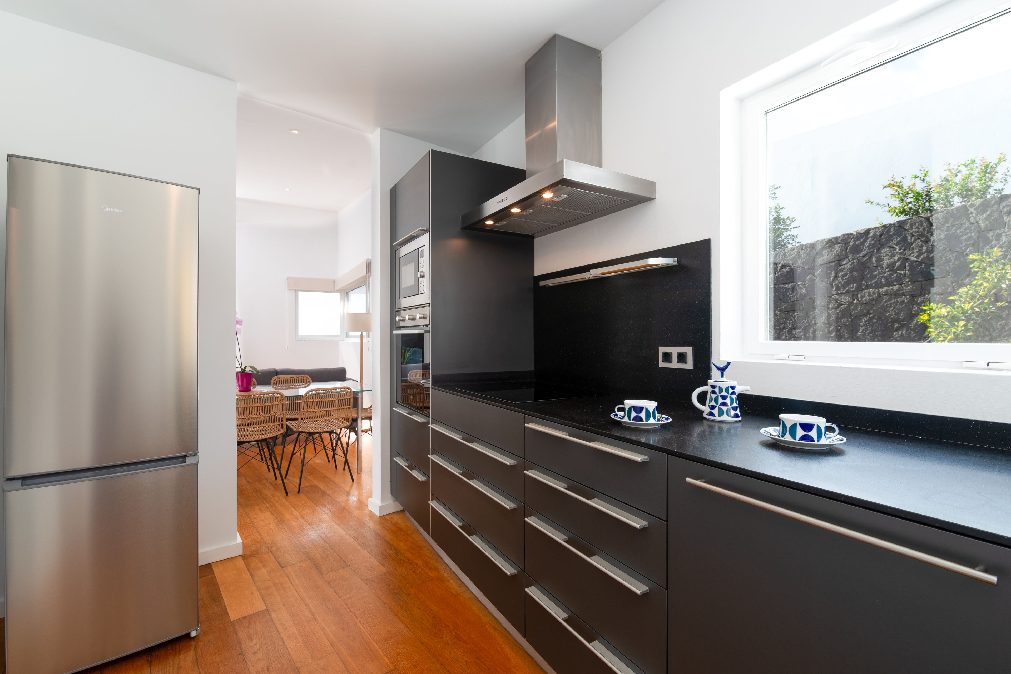 A modern kitchen with stainless steel appliances, a black countertop, and a window overlooking a stone wall.