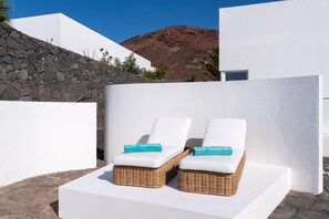Zwei Korbsessel mit weißen Kissen und blauen Handtüchern, platziert auf einer weißen Plattform mit einer Steinmauer und einem Berg im Hintergrund.