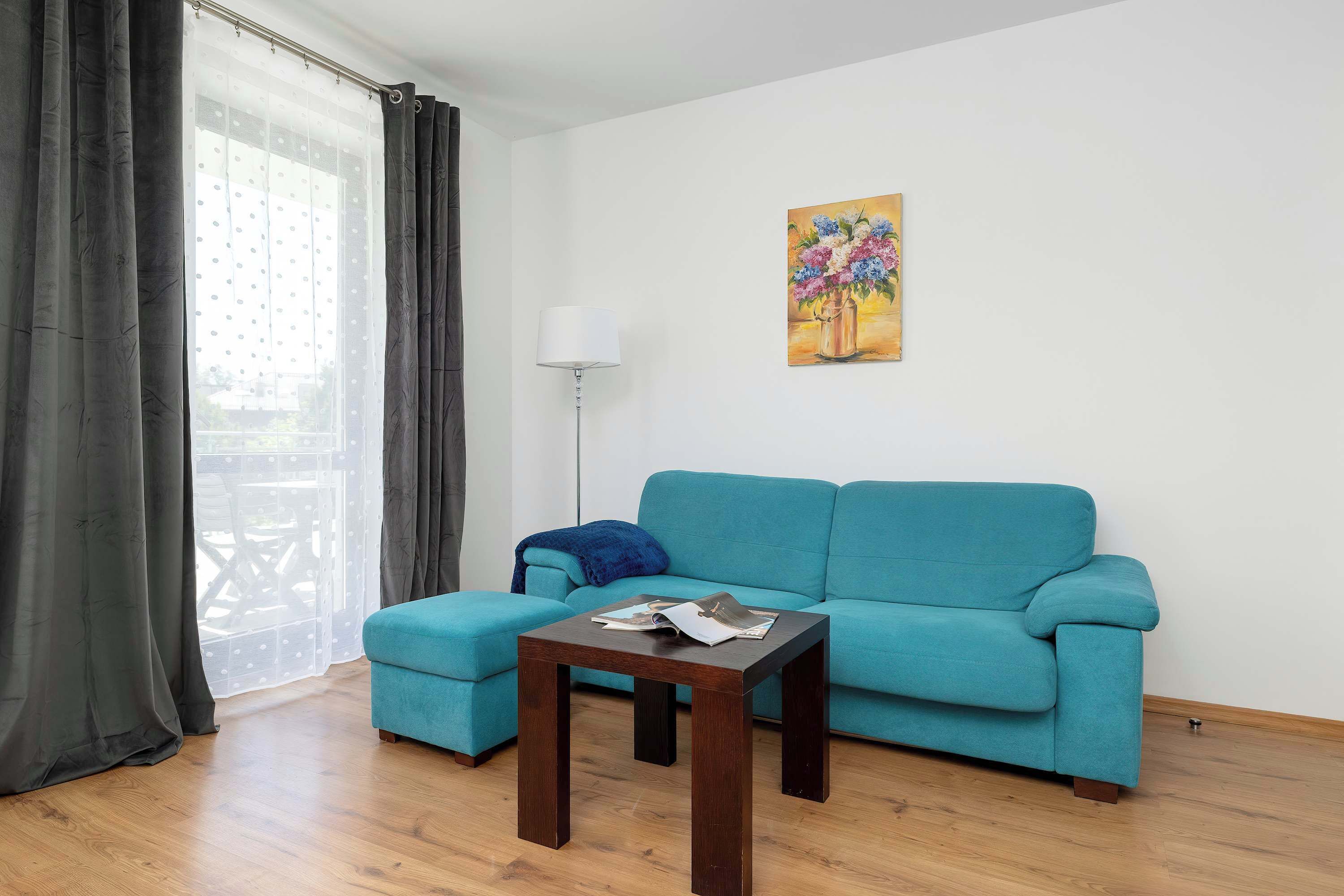 A living room with a teal sofa, a small wooden table, a floor lamp, and a painting on the wall.