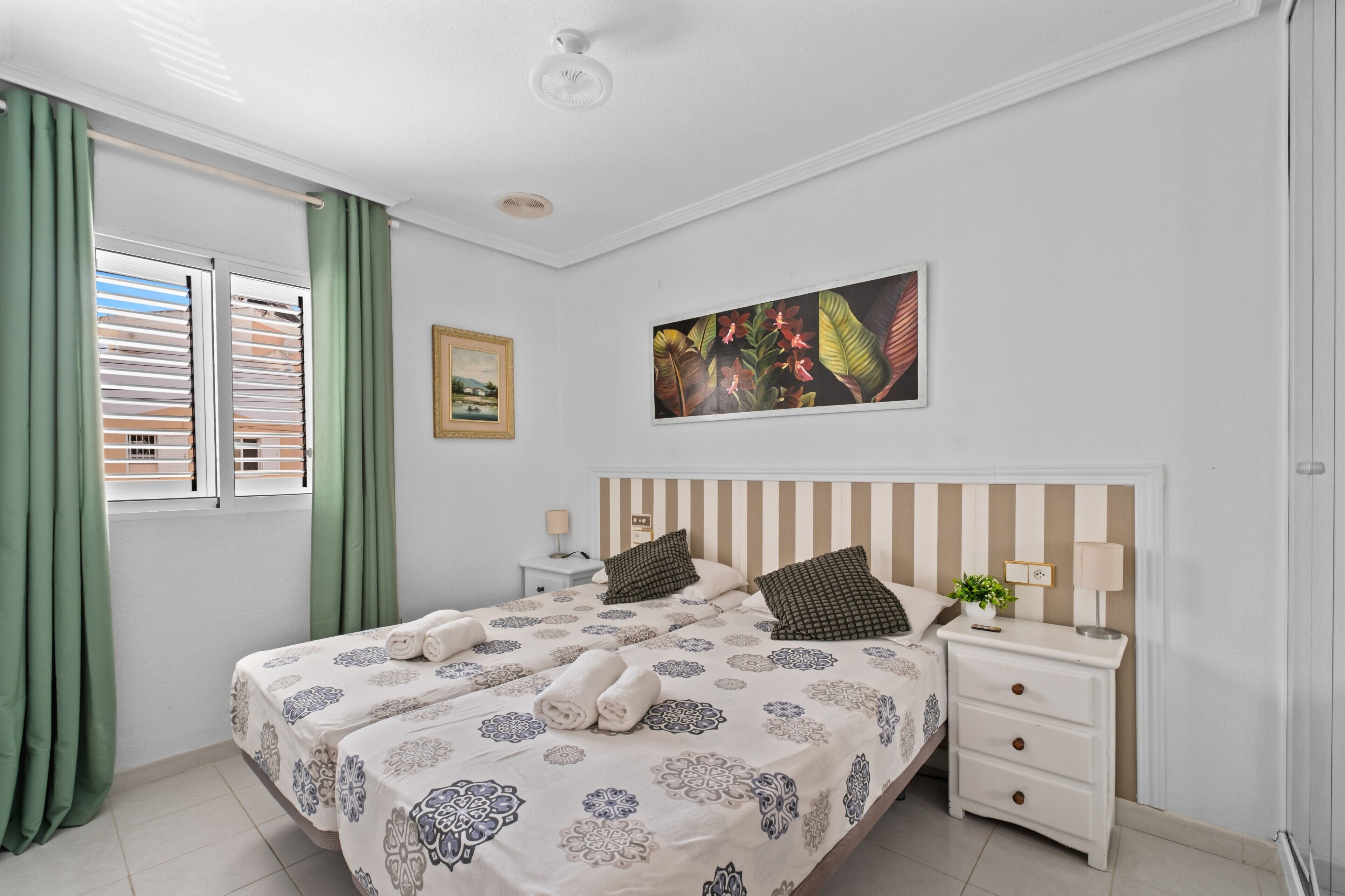 A bedroom with two beds, a nightstand, a wall-mounted TV, and a window with curtains.