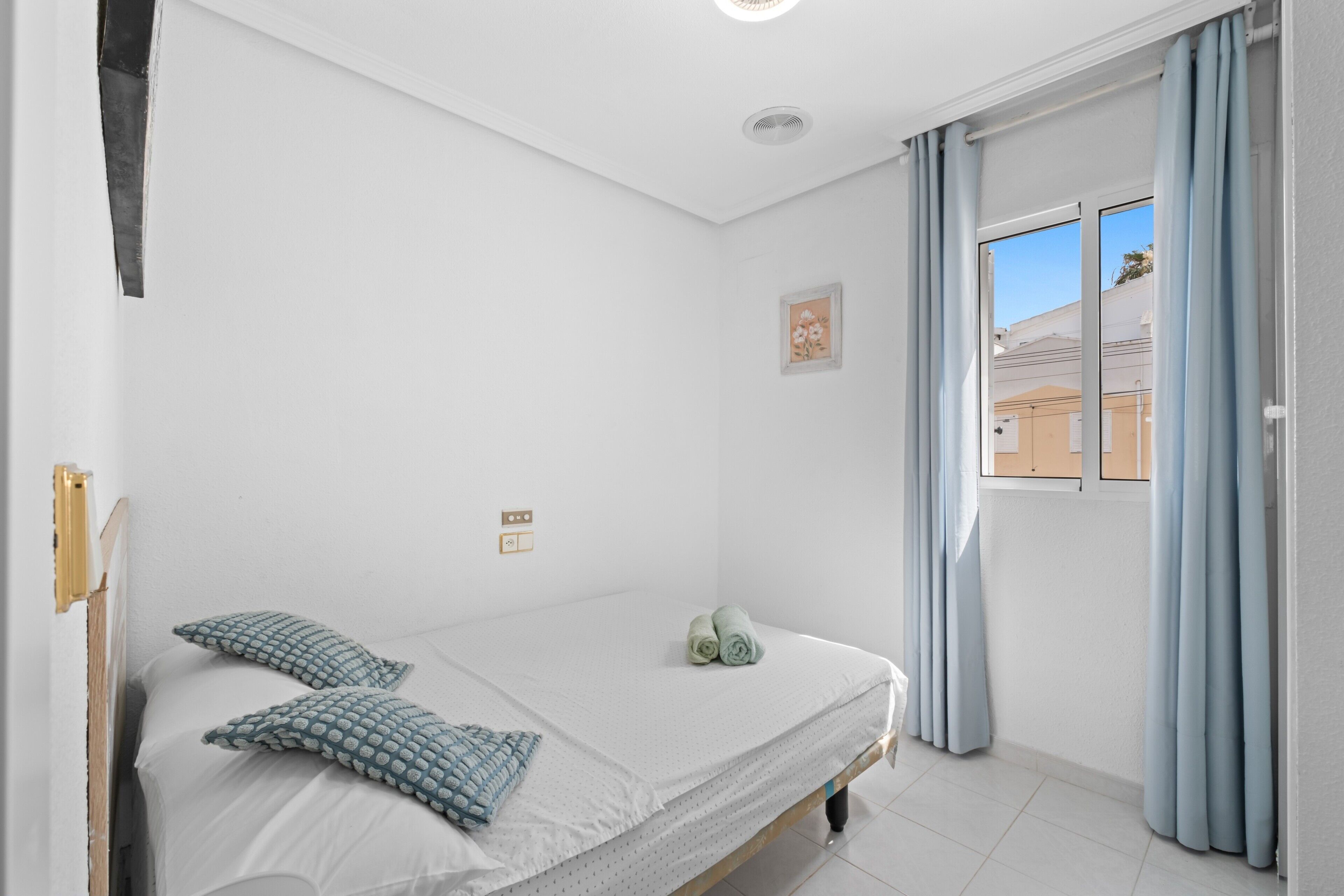 Een slaapkamer met een bed, kussens, een klein schilderij en een raam met gordijnen.