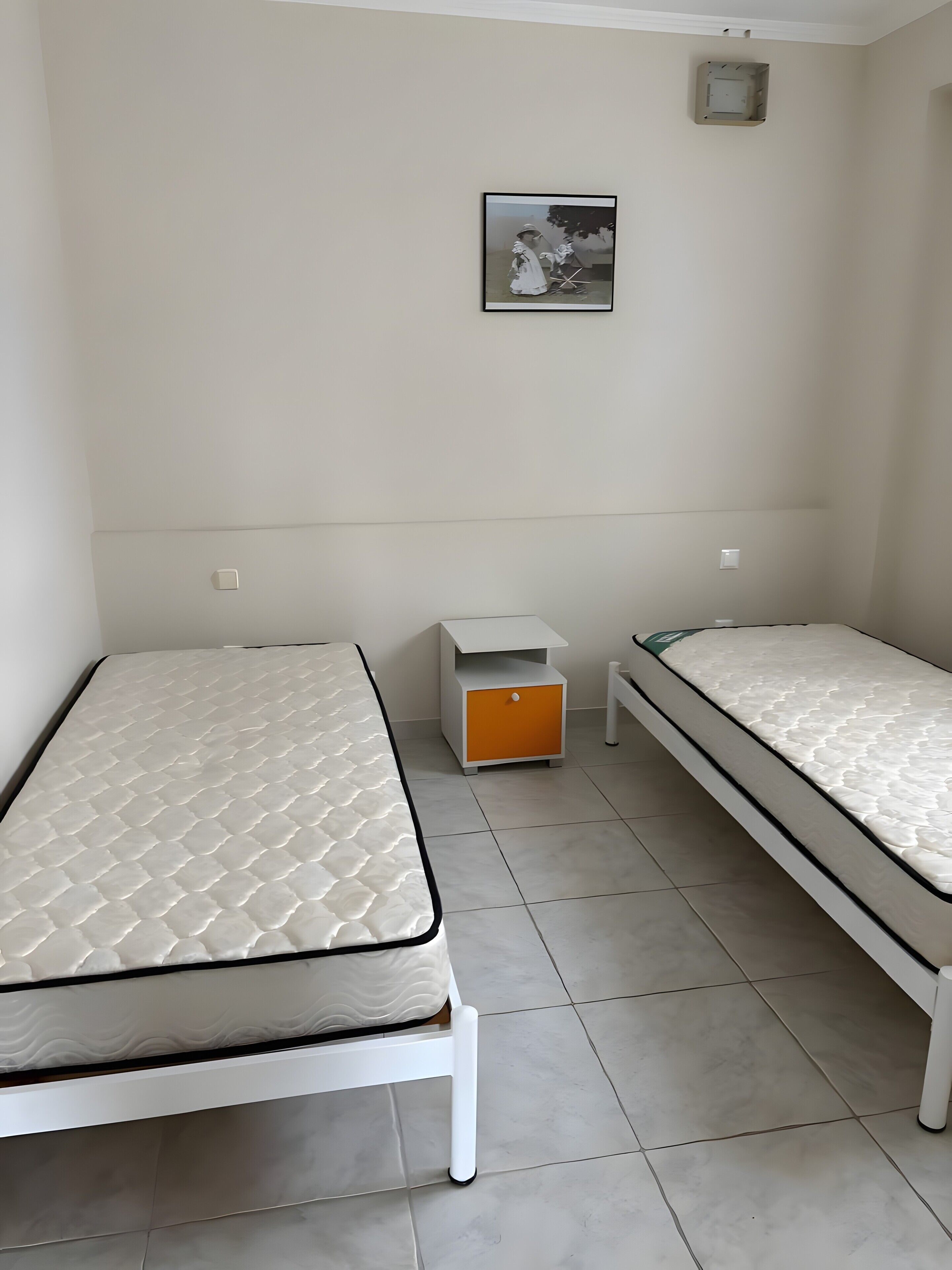 Una habitación con dos camas individuales, una mesita de noche pequeña de color blanco con un cajón naranja, un cuadro en la pared y un respiradero blanco en el techo.