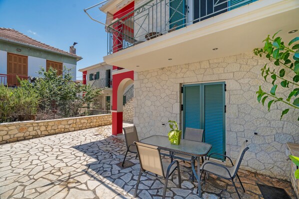 A stone-floored patio with a table and chairs, a door with blue shutters, and a balcony with red railings.