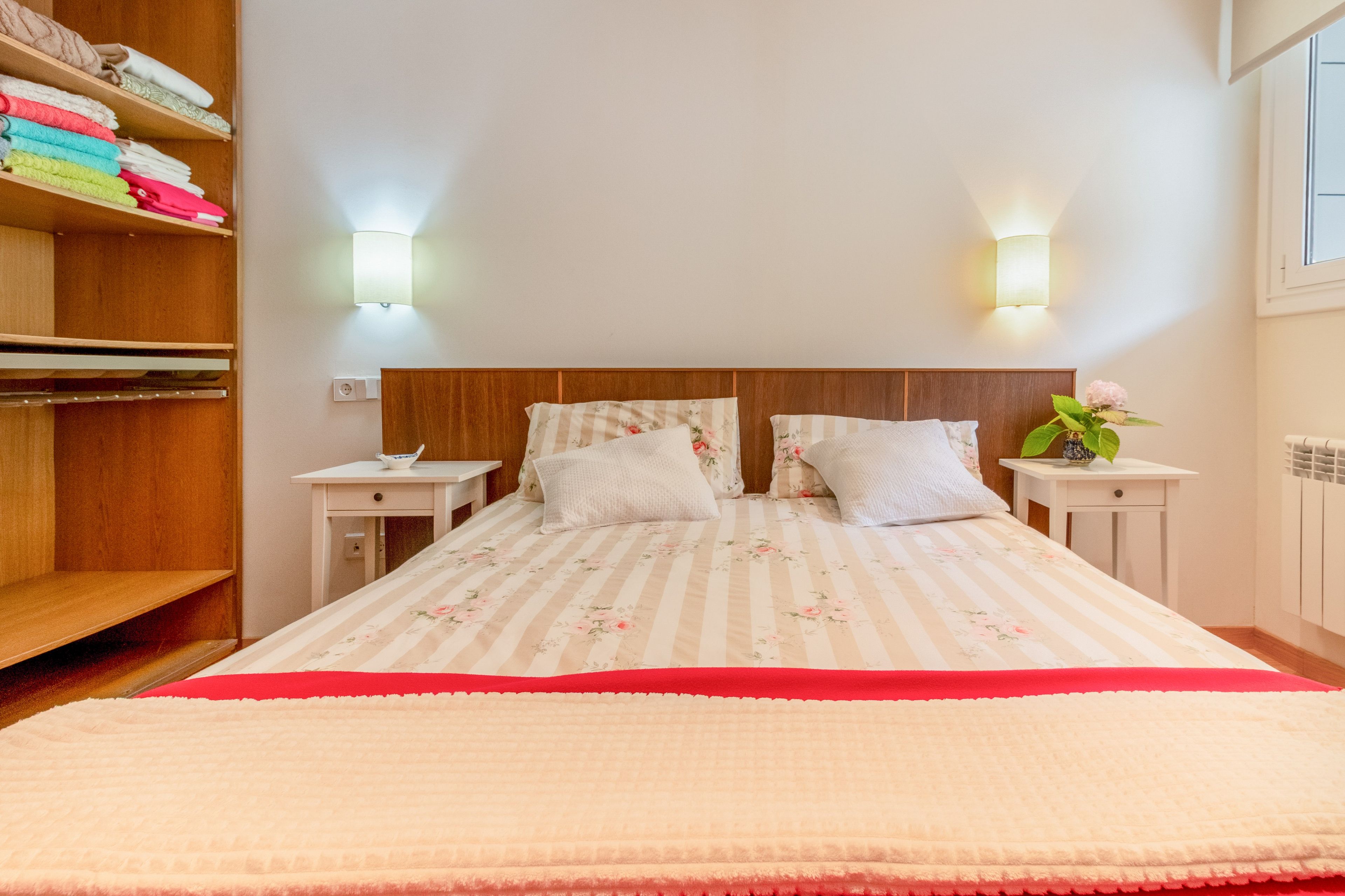Una cama bien hecha con ropa de cama floral, una mesita de noche con una lámpara y un estante con toallas dobladas.
