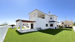 Una casa moderna blanca con un amplio jardín, una pérgola y un cielo azul despejado.