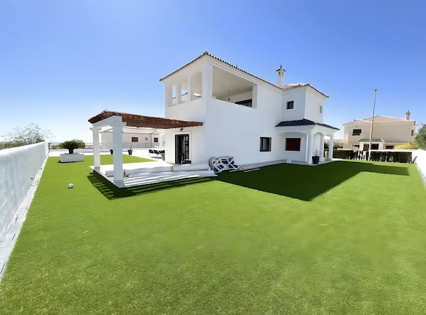 Une maison moderne aux murs blancs, dotée d’une grande pelouse, d’une pergola et surplombée d’un ciel d’un bleu éclatant.