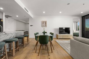 A modern living room with a TV, a dining area with a table and chairs, a kitchen with a marble countertop, and a large window. - Luxurious 2BR In Sought-After `Eastbourne` Complex (Parkes)
