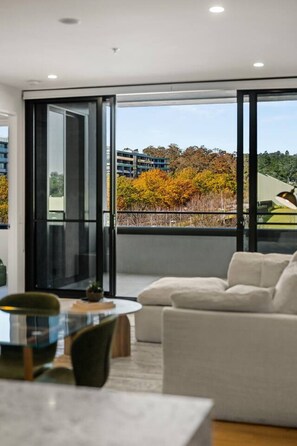 Ein modernes Wohnzimmer mit einer Couch, einem Esstisch und Blick auf ein Gebäude und Bäume durch große Fenster.