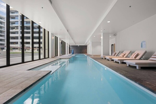A modern indoor swimming pool with clear glass walls, a long pool, and lounge chairs along the right side. - Luxurious 2BR In Sought-After `Eastbourne` Complex (Parkes)