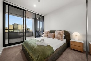 A modern bedroom with a large sliding glass door leading to a balcony, a neatly made bed, a bedside table with a lamp, and a view of buildings. - Luxurious 2BR In Sought-After `Eastbourne` Complex (Parkes)