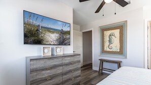 Une chambre à coucher comprenant un lit, une commode, un tableau représentant une scène de plage, un tableau représentant un hippocampe et un ventilateur de plafond.