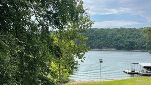 Une zone herbeuse avec un quai et un bateau, un lac, et une colline boisée en arrière-plan.