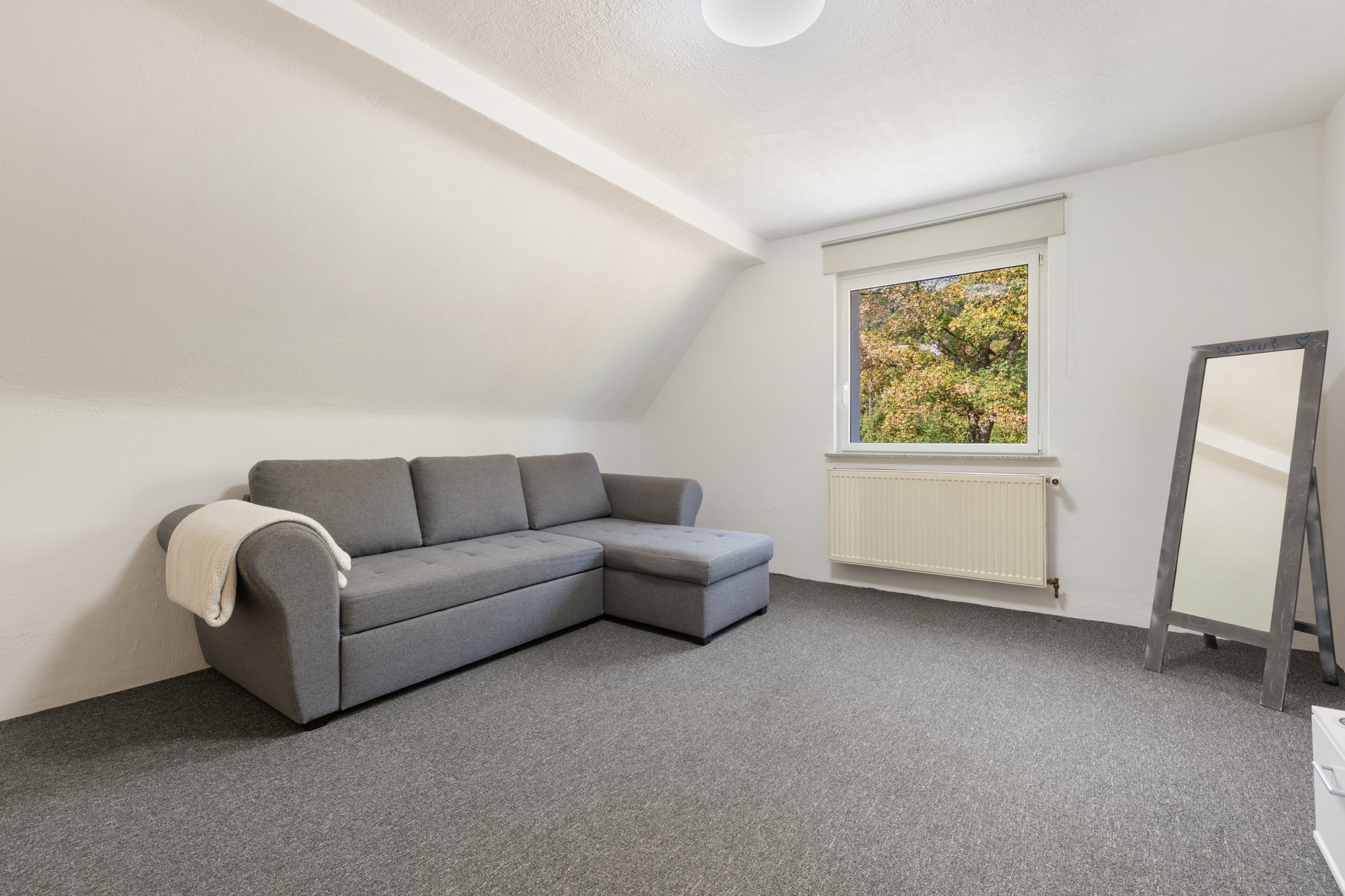 A room with a grey sectional sofa, a radiator, and a window with a view of trees.