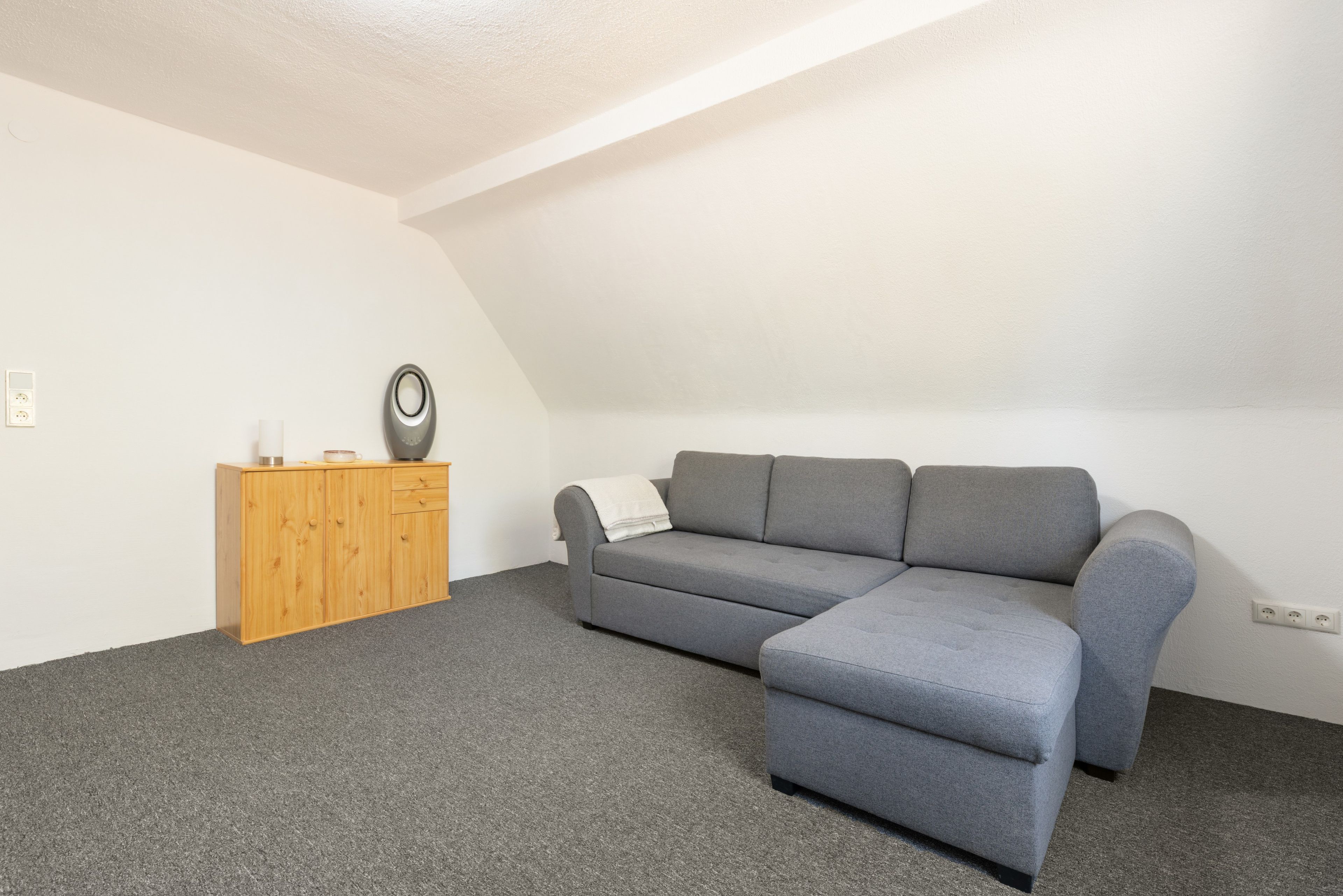 Une pièce comprenant un canapé d’angle gris, un meuble en bois et une petite table avec une bouilloire.