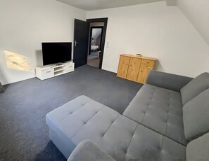 Ein Wohnzimmer mit grauem Sofa, einem Flachbildfernseher, einem Holzschrank und einer Tür, die in einen anderen Raum führt.