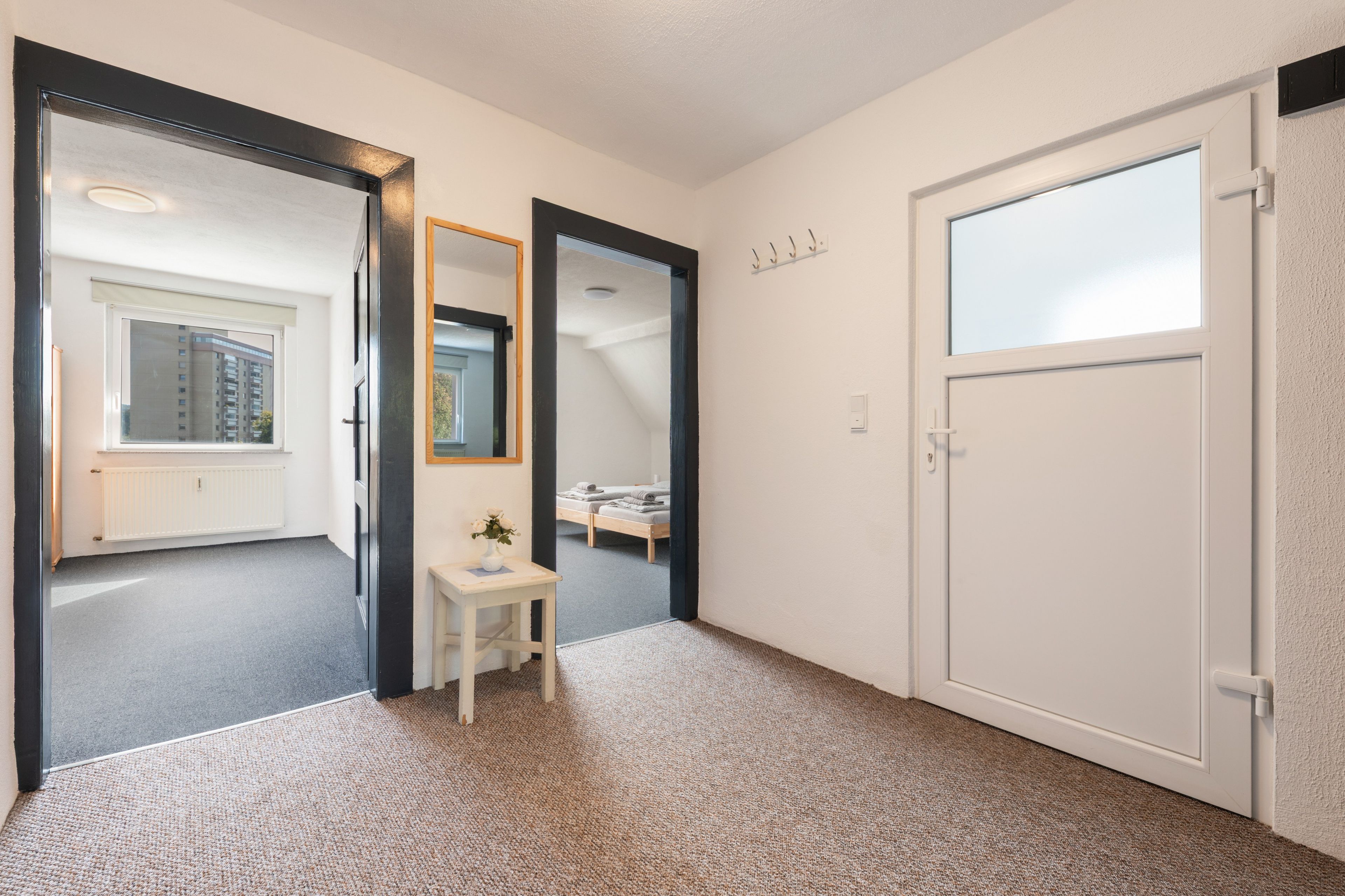 Un pasillo con una puerta blanca, un espejo, una mesita con un florero y vistas a un dormitorio con cama y ventana.