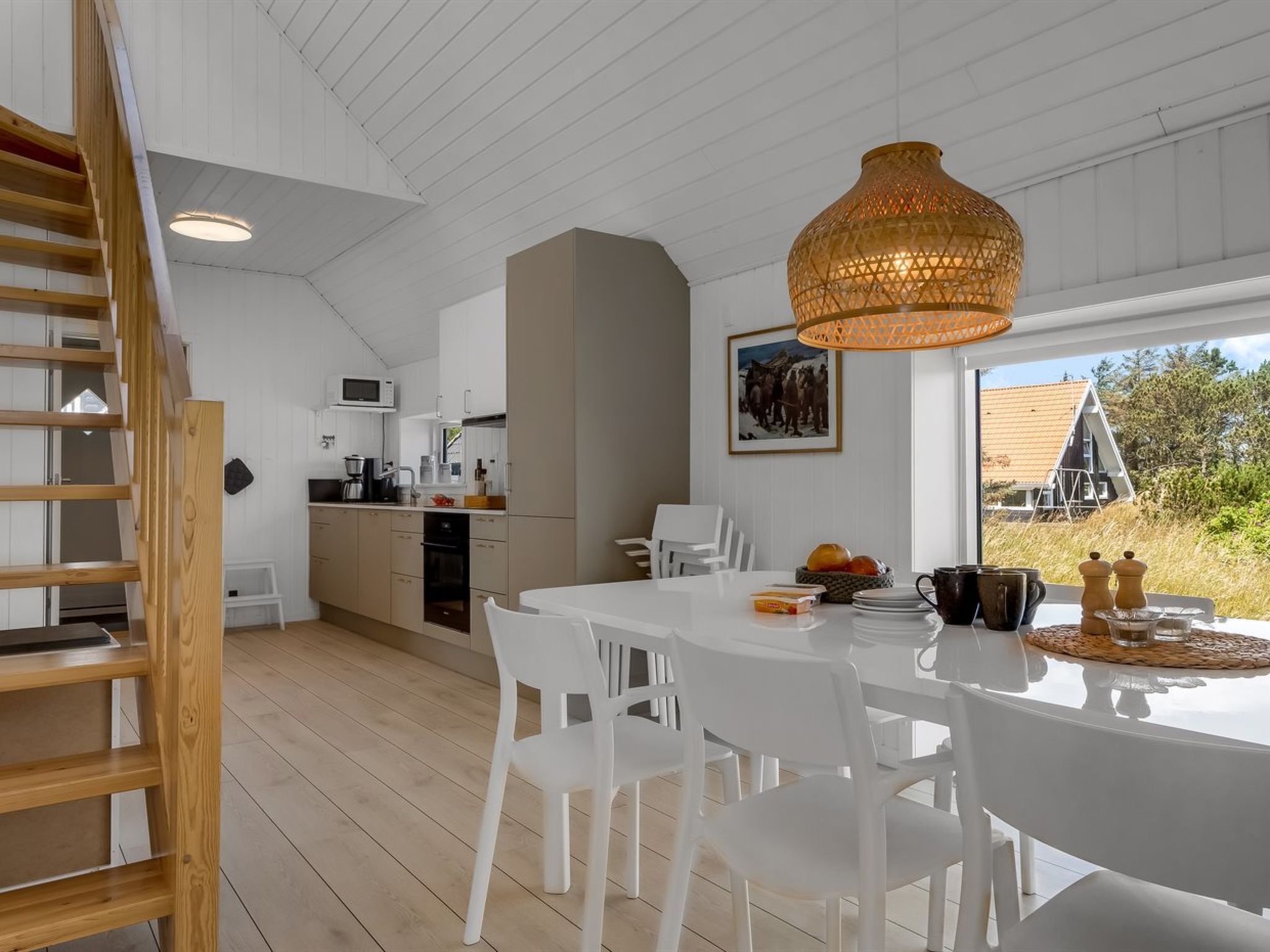 A modern kitchen with white cabinets, a dining table with chairs, a hanging light fixture, and a view of the outside.