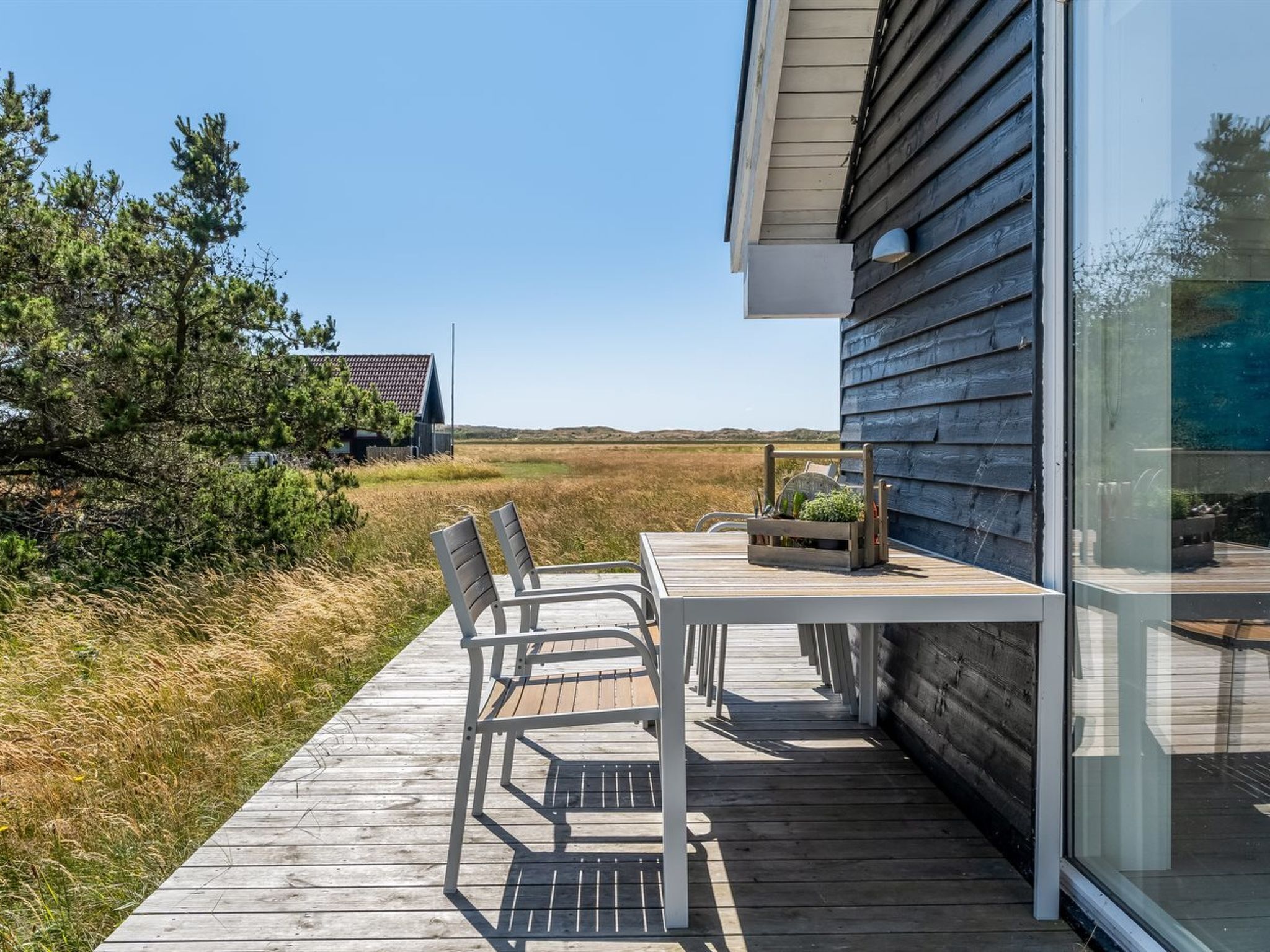 Une terrasse en bois avec une table et des chaises, une porte vitrée et une vue sur une maison et le paysage environnant.