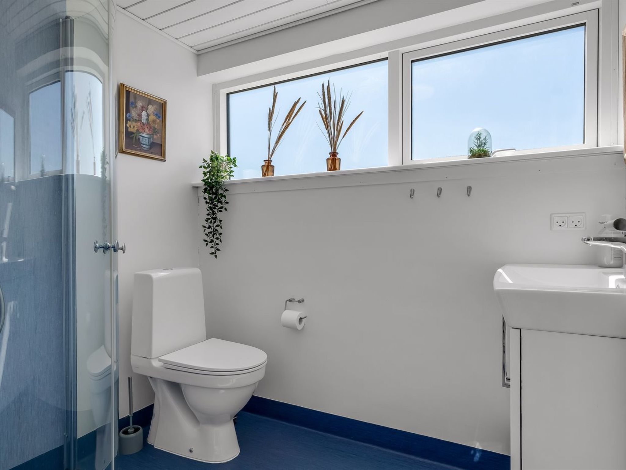 Un baño con inodoro, lavamanos, ventana con plantas y zona de ducha.