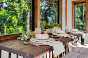 A dining table set with white plates, bowls, and cutlery, with a view of greenery outside.