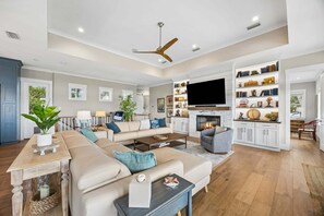 A spacious living room with a large sofa, a coffee table, and a fireplace. There is a television mounted above the fireplace and a ceiling fan. - Oyster Manor|Marsh Views|Heated Pool+Spa|Vilano Beach (St. Augustine)