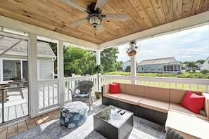 A covered porch with a ceiling fan, a cushioned seating area, and a view of a house and greenery.