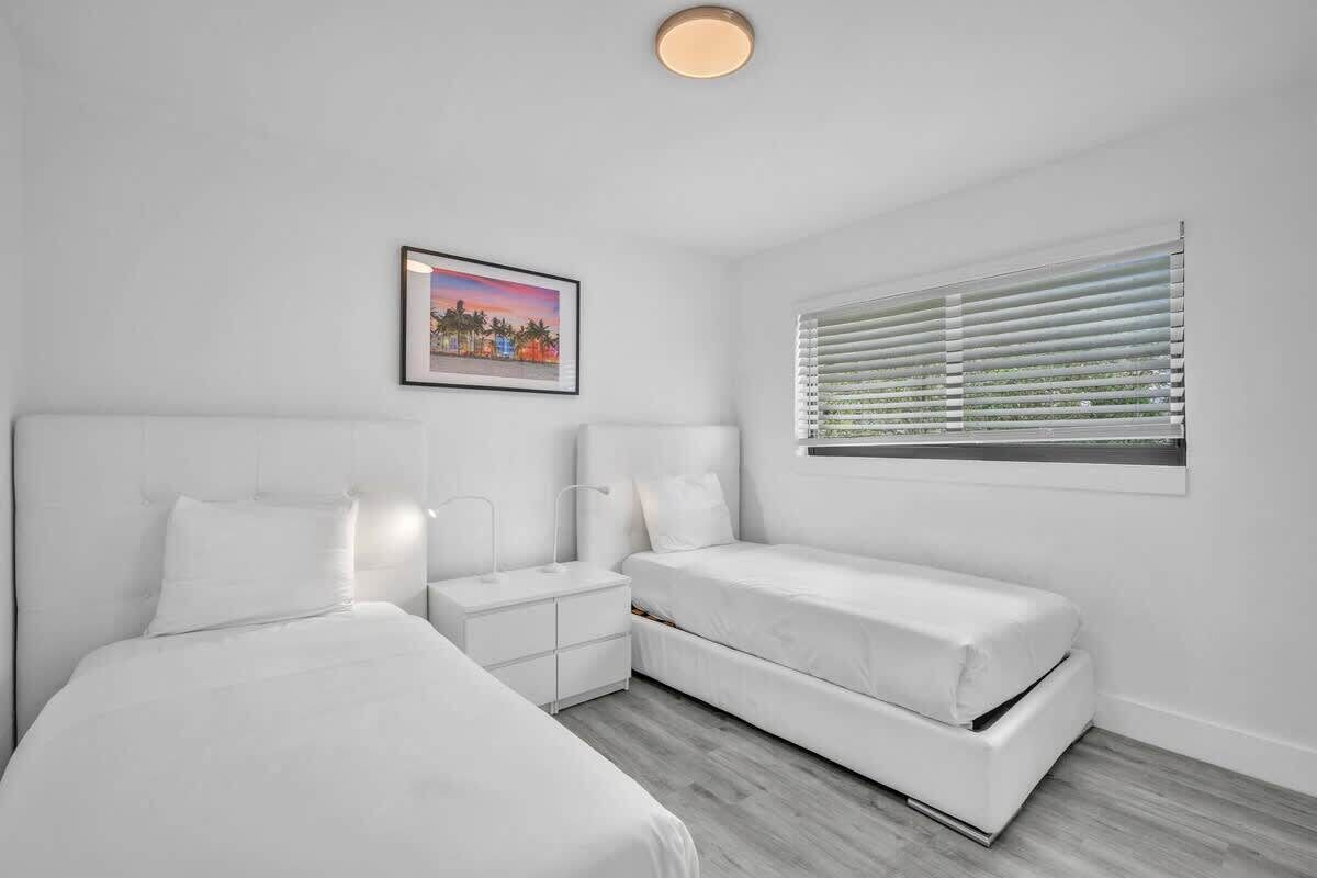 A modern bedroom with two beds, a nightstand, a framed picture, and a window with blinds.