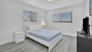 Een moderne slaapkamer met een wit bed, twee nachtkastjes, een televisie en ramen met zonwering.