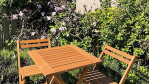 Ein Holztisch mit Stühlen steht auf einer Terrasse, im Hintergrund eine Backsteinmauer und viel Grün.