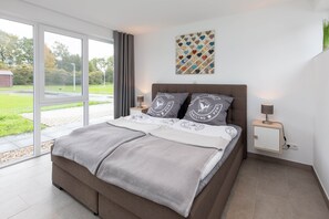Ein modernes Schlafzimmer mit einem großen Bett, einer Schiebetür zu einem Garten, einem kleinen Nachttisch mit Lampe und einem an der Wand befestigten Schrank.