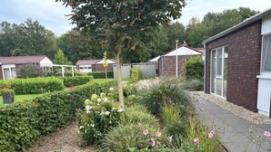 Un jardín con setos, plantas en flor y un sendero que conduce a una hilera de edificios de ladrillo con ventanas.