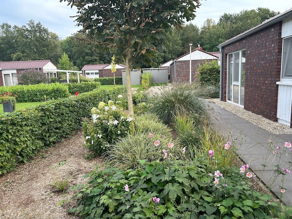 Un jardín con setos, plantas en flor y un sendero que conduce a una hilera de edificios de ladrillo con ventanas.