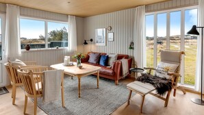A cozy living room with a wooden ceiling, a brown leather sofa, a dining table with chairs, and large windows offering a view of the outdoors. - "Sida" - 400m from the sea by Interhome (Løkken)