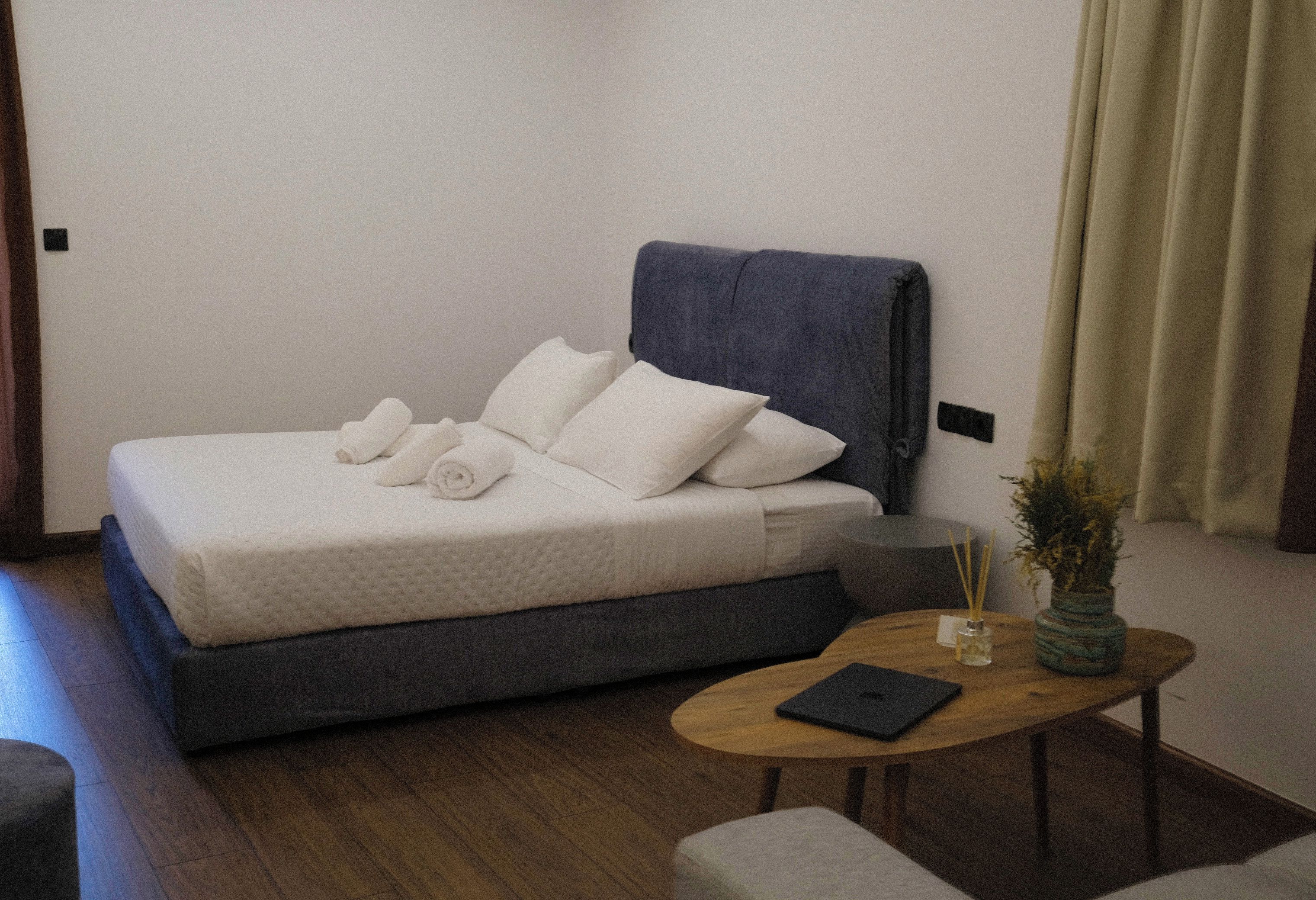 A neatly made bed with pillows and a rolled towel, a bedside table with a book and a vase, and a small round table with a candle and reed diffuser.